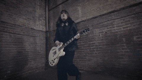 a woman in a black jacket is playing a white guitar in front of a brick wall