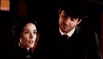 a man and a woman are standing next to each other in a dark room . the man is wearing a top hat .