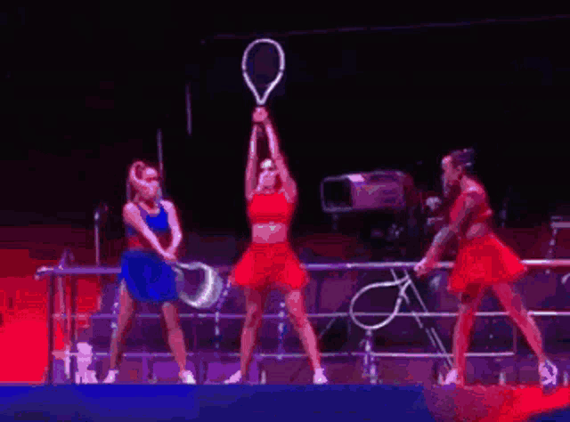 three women holding tennis rackets on a stage with a camera in the background