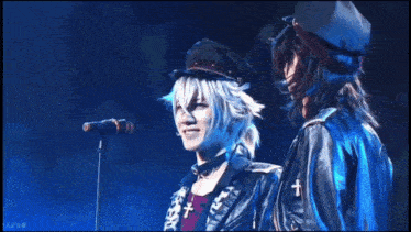 a man singing into a microphone next to another man with a cross on his jacket