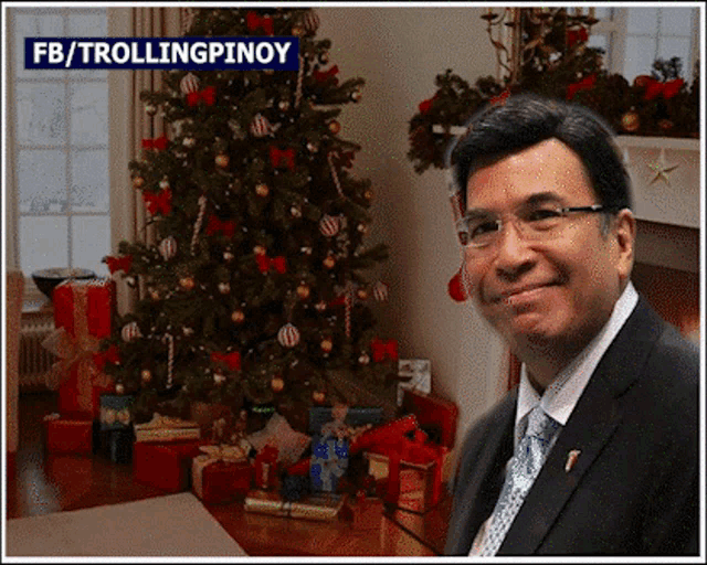 a man in a suit and tie stands in front of a christmas tree with gifts under it