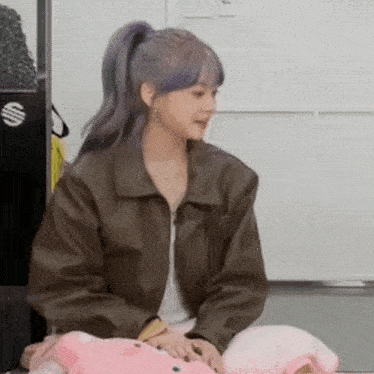 a woman in a brown leather jacket is sitting on a couch holding a pink pillow .