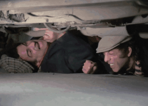 two men are laying under a car and one is wearing a flat cap