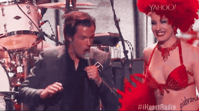 a man is talking into a microphone while a woman in a red bra stands behind him with yahoo in the background