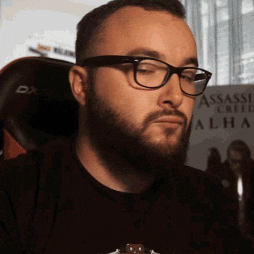 a man with glasses and a beard is sitting in front of a assassin creed poster