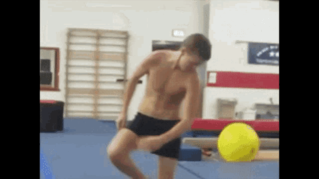 a man without a shirt is doing a trick with a yellow ball in a gym .