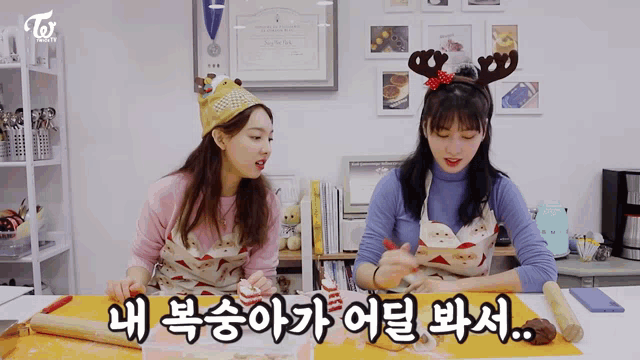 two women wearing aprons and reindeer antlers sit at a table with a rolling pin