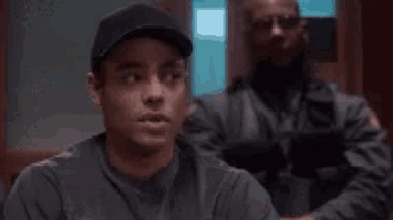 a man in a baseball cap is sitting in a room with a police officer standing behind him .