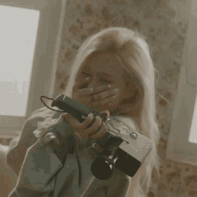 a woman with blonde hair is holding a camera and covering her mouth with her hand .