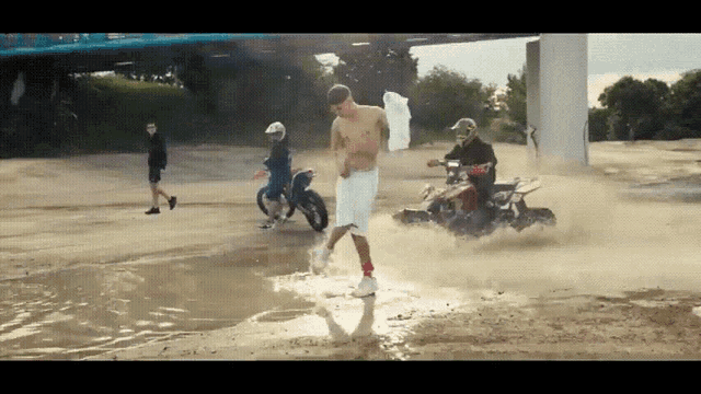 a man without a shirt is walking through a puddle of water while a man on a motorcycle rides by