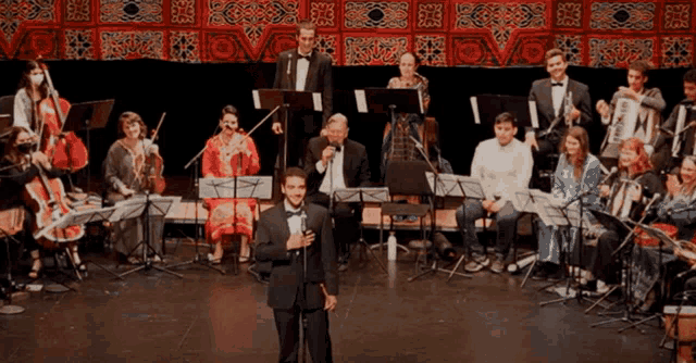 a man in a tuxedo sings in front of a large orchestra