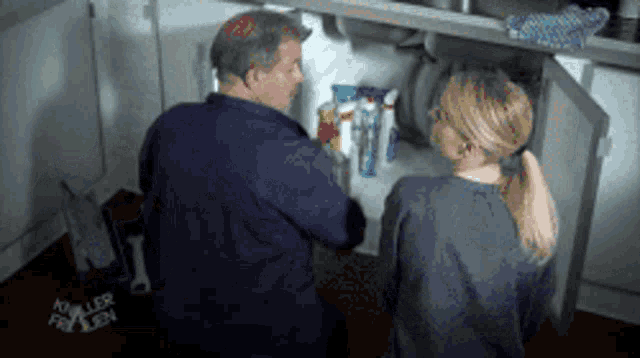a man and a woman are looking under a sink and the man has the word killer on his shirt