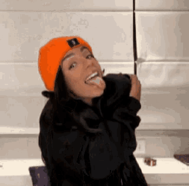 a woman wearing an orange beanie and a black jacket is sticking out her tongue .