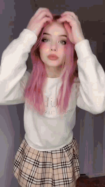 a woman with pink hair is wearing a plaid skirt and a white sweater .