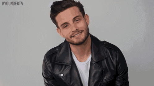 a man with a beard wearing a black leather jacket and a white shirt is smiling .