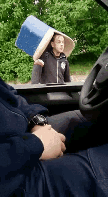 a man is carrying a bucket on his head while sitting in a car