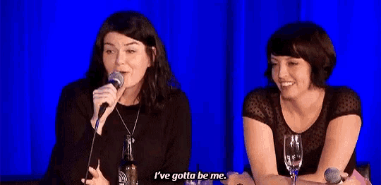 two women are sitting at a table and one is holding a microphone and the other is saying i 've gotta be me