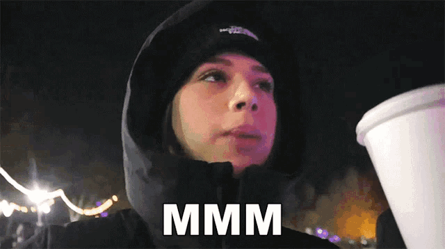 a woman wearing a hooded jacket is holding a cup and the word mmmm is above her