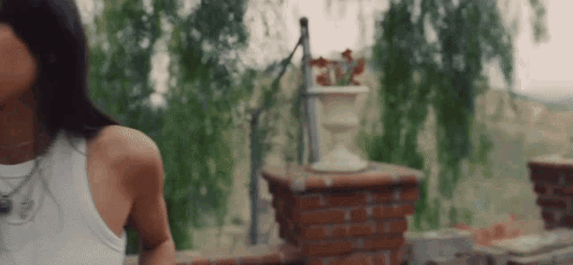 a woman in a white tank top is standing in front of a brick wall with a vase in the background .