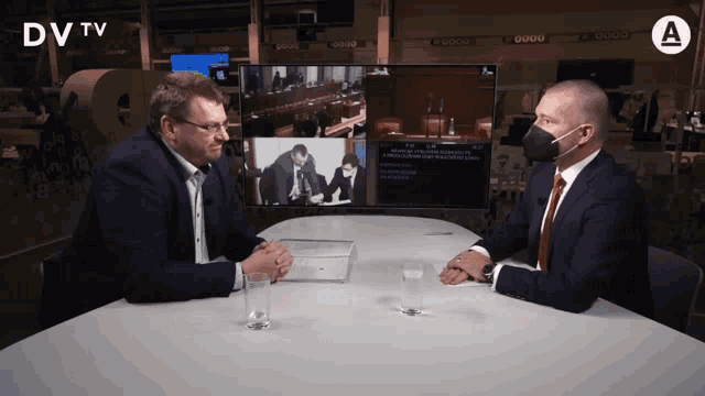 two men are sitting at a table talking in front of a dv tv screen
