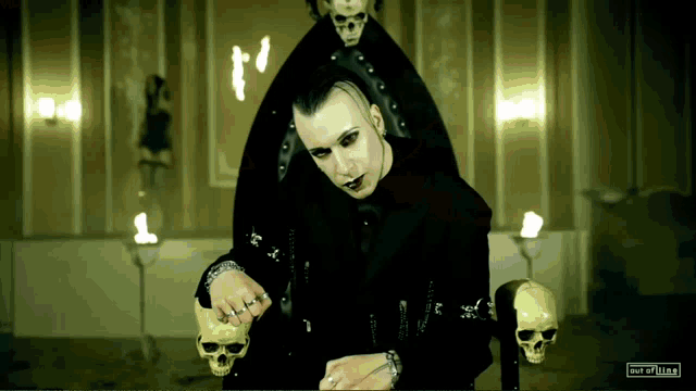 a man in a black suit sits in a chair with skulls on the back