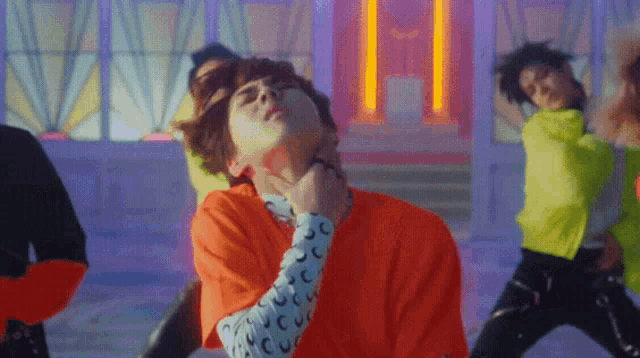 a man in an orange shirt is dancing in front of a neon sign that says h