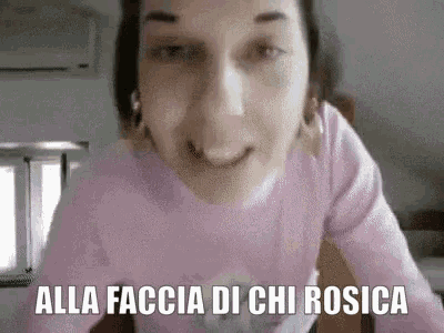 a woman in a pink sweater is making a funny face with the words `` alla faccia di chi rosica '' .