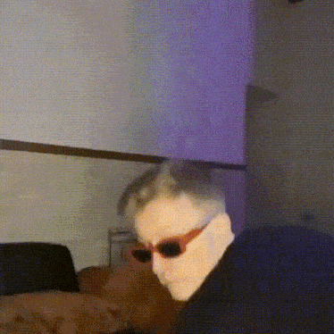 a man wearing sunglasses is sitting on a couch in front of a purple wall .