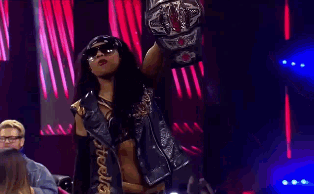 a woman wearing sunglasses and a bandana is holding a championship belt on a stage .