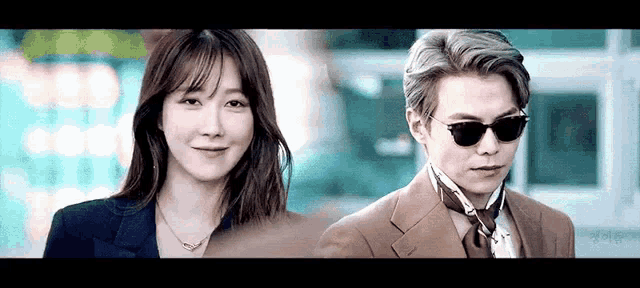 a man and a woman are standing next to each other . the man is wearing sunglasses and a suit .