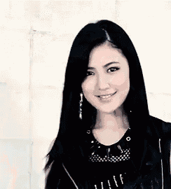 a woman with long black hair is standing in front of a white wall wearing a black vest and earrings .