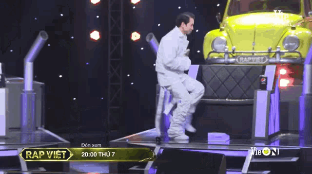 a man in a white suit is sitting on a stool on a stage in front of a yellow car .