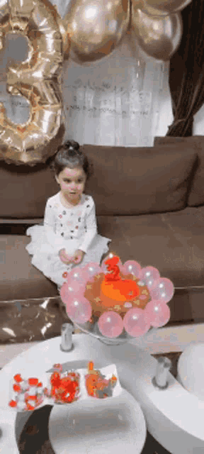 a little girl is sitting on a couch with balloons and a cake in front of her