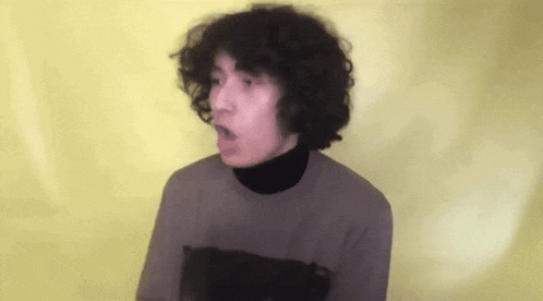 a young man with curly hair is wearing a turtleneck and a sweater and making a funny face .