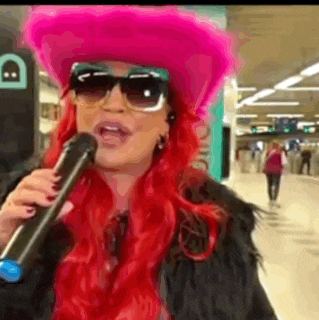 a woman with red hair and sunglasses is singing into a microphone while wearing a pink hat .