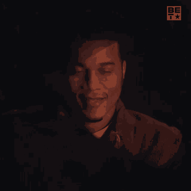 a man is smiling in a dark room with a be t logo in the corner