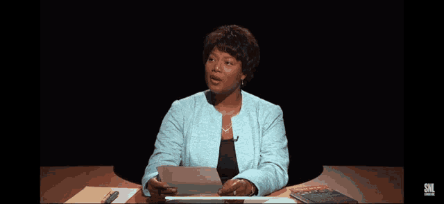 a woman sitting at a desk holding a piece of paper with a snl logo on the bottom