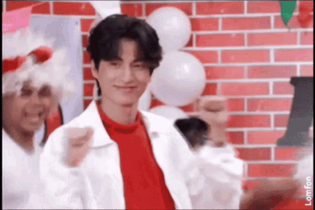 a man in a red shirt and white jacket is standing in front of a brick wall with balloons .