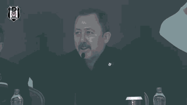 a man with a mustache is talking into a microphone while sitting at a table with bottles of water .
