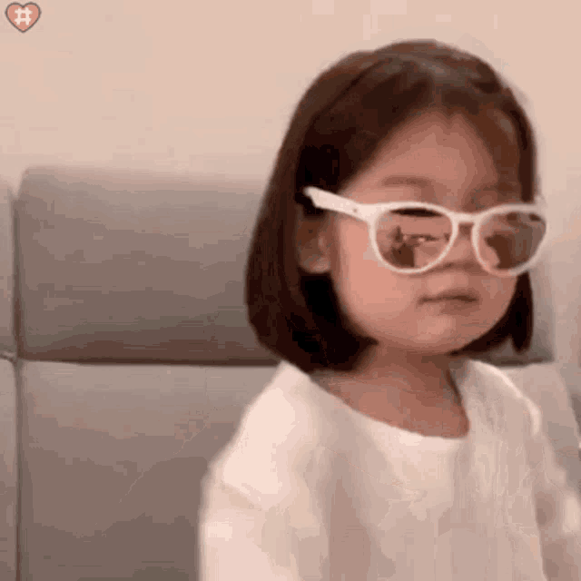 a little girl wearing sunglasses is sitting on a couch looking at the camera .