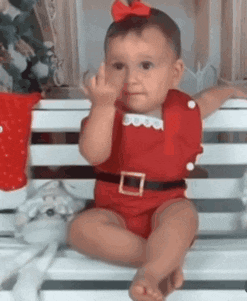 a baby dressed in a santa suit is sitting on a bench giving the middle finger .