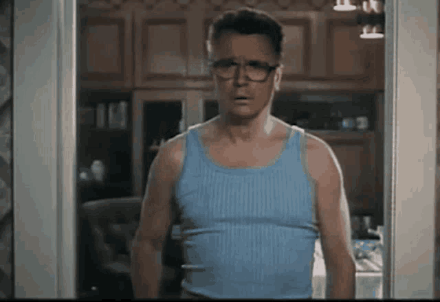 a man in a blue tank top and glasses is standing in a kitchen