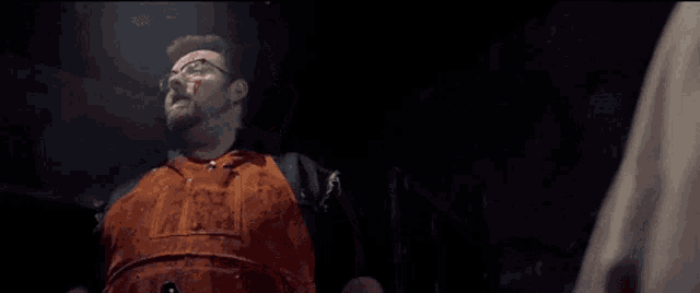 a man with glasses and an apron is standing in a dark room with blood on his shirt .
