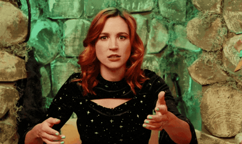 a woman with red hair and green nails is standing in front of a stone wall with her hands outstretched