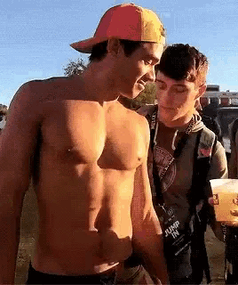 a man without a shirt is standing next to another man who is holding a cup of beer .