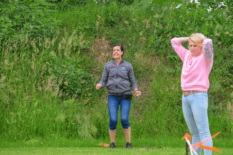 a woman in a pink sweater applauds while another woman in a grey jacket stands in the grass
