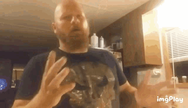 a bald man with a beard wearing a black shirt is standing in a kitchen making a funny face .