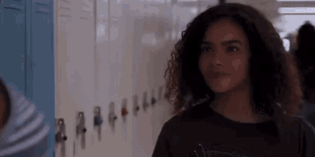 a girl with curly hair is standing in front of a row of lockers in a hallway .