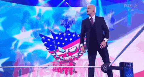 a man in a suit and tie is standing in a wrestling ring with a flag in the background .