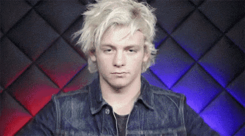 a man with blonde hair and a denim jacket is making a funny face .
